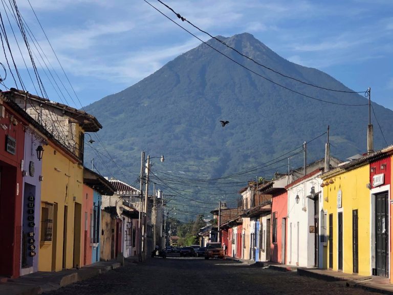 The main colorful street of antigua guatemala. Bright wooden buildings frame a cobblestone street. A large volcano is seen in the distance.